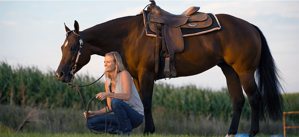Woman kneeling outside in a pasture next to a brown horse's front legs with a saddle at sunset